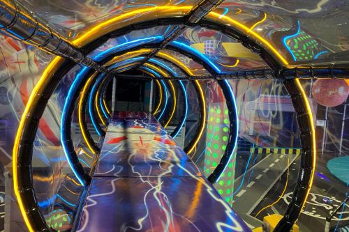 A colorful, illuminated tunnel with swirling patterns and bright lights, leading to a playful, vibrant space.