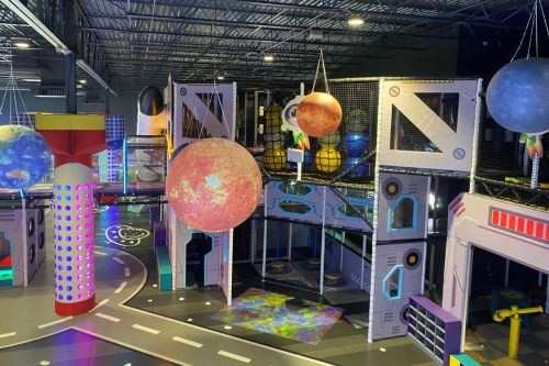 A colorful indoor play area featuring large hanging planets, space-themed decorations, and vibrant lighting.