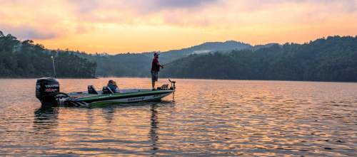 A person fishing from a boat on a calm lake at sunrise, surrounded by hills and misty scenery.