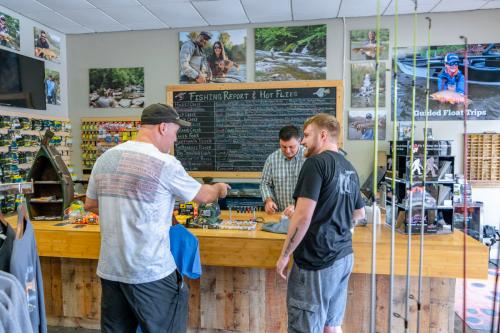 Two customers interact with a staff member at a fishing shop, surrounded by fishing gear and a chalkboard report.