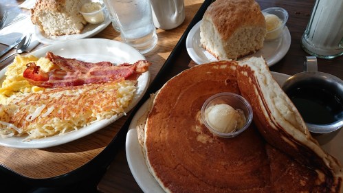 A breakfast spread featuring pancakes with butter, bacon, scrambled eggs, hash browns, and a biscuit with butter.