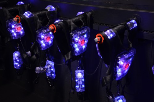 A row of futuristic laser tag vests and guns with glowing blue and red lights, hanging on a dark wall.