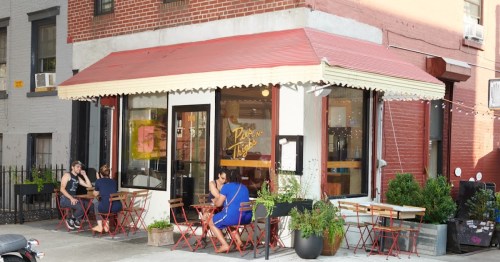 A cozy café with outdoor seating, featuring red chairs and a bright awning, surrounded by brick buildings.