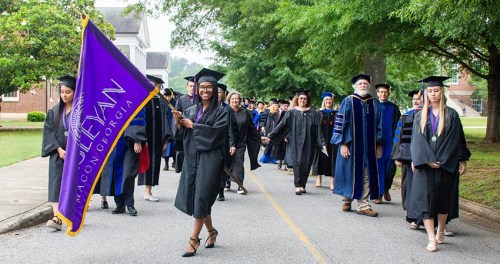 A group of graduates in caps and gowns walking together, with one holding a purple flag, on a tree-lined path.