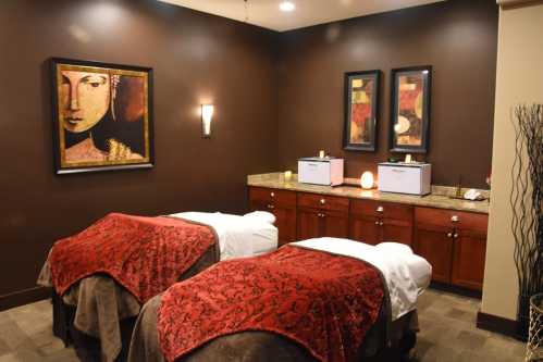 A serene spa room with two massage tables, adorned with red covers, and calming artwork on the walls.