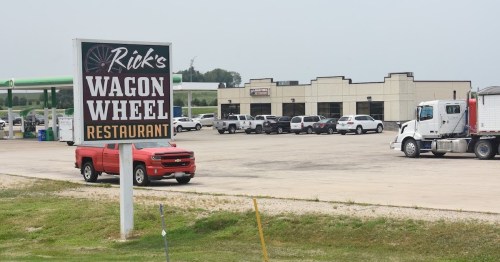 Sign for Rick's Wagon Wheel Restaurant with a red truck parked nearby and a view of a parking lot and buildings.