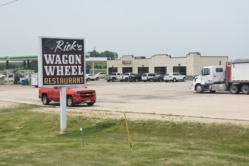 Sign for Rick's Wagon Wheel Restaurant with a red truck parked nearby and a truck in the lot, surrounded by a grassy area.