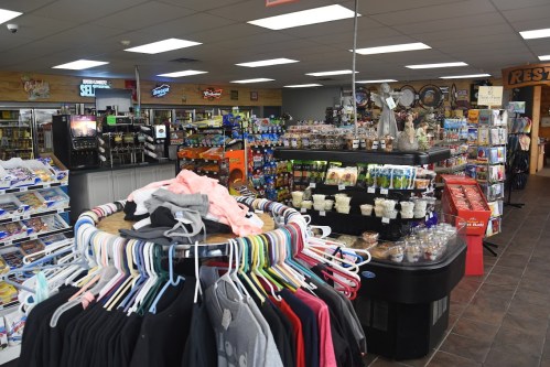 A convenience store interior featuring clothing racks, snacks, drinks, and various merchandise displays.