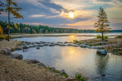A serene lakeside scene at sunset, with rocks, trees, and a colorful sky reflecting on the water's surface.
