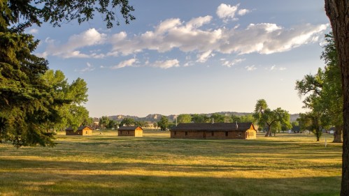 A serene landscape featuring several wooden buildings surrounded by green grass and trees under a partly cloudy sky.
