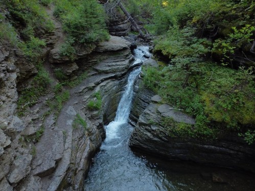 A serene waterfall cascades through rocky cliffs, surrounded by lush greenery and trees in a tranquil natural setting.