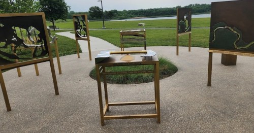 A circular outdoor installation with gold-framed art pieces and a central table, surrounded by grass and a lake.