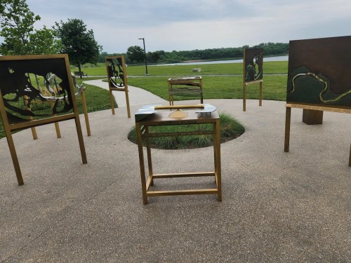 A circular arrangement of gold-framed art pieces and a table in a grassy park by a lake under a cloudy sky.