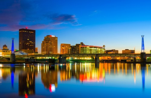 City skyline at dusk, reflecting in the water, with illuminated buildings and a bridge under a blue sky.