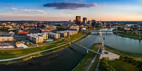 Aerial view of a city skyline at sunset, featuring buildings, a river, and a pedestrian bridge.