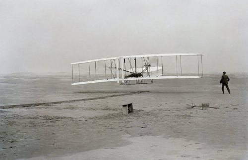 Historic image of the Wright brothers' first powered flight, with the aircraft taking off and a man observing nearby.