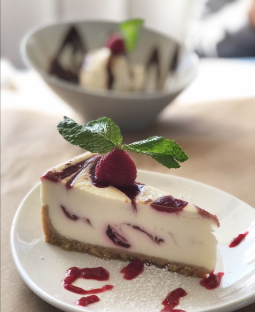 A slice of cheesecake topped with raspberry sauce and a mint leaf, served on a white plate with a blurred dessert in the background.