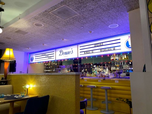 A brightly lit bar area with a sign reading "Denae's Diner," featuring colorful lights and a variety of drinks on display.