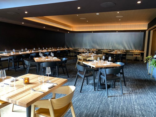 A modern restaurant interior featuring wooden tables, black chairs, and a minimalist design, set for dining.
