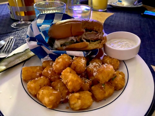 A plate with a sandwich in a blue checkered wrapper, tater tots, a pickle, and a small bowl of dipping sauce.