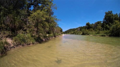 A serene river scene with clear water, sandy banks, and lush greenery under a bright blue sky.