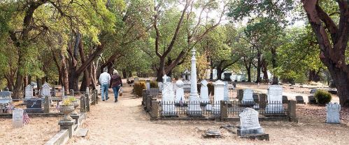 Two people walk through a serene cemetery surrounded by trees and gravestones.