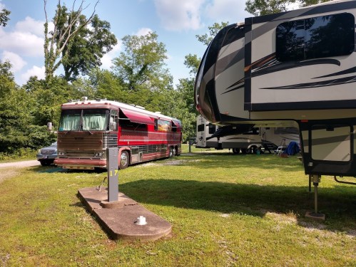 A red and silver RV parked next to a large fifth-wheel trailer in a grassy campground surrounded by trees.