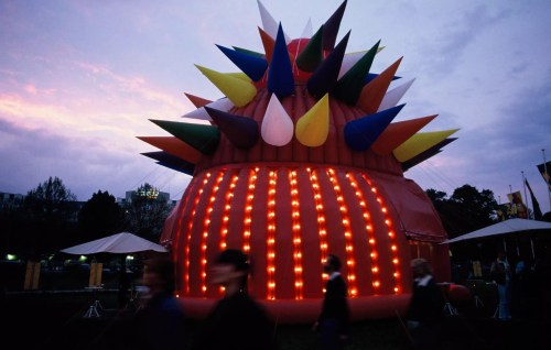 A large, colorful inflatable structure with spikes and lights, set against a twilight sky with people walking by.