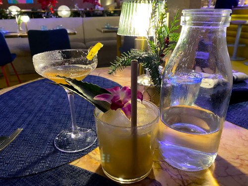 A table setting featuring two cocktails, one garnished with an orchid, and a glass bottle of water.