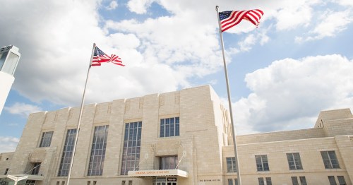 A stone building with large windows, featuring two American flags flying in front against a partly cloudy sky.