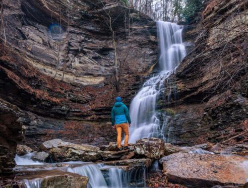 A person in a teal jacket stands near a waterfall cascading down rocky cliffs, surrounded by autumn foliage.