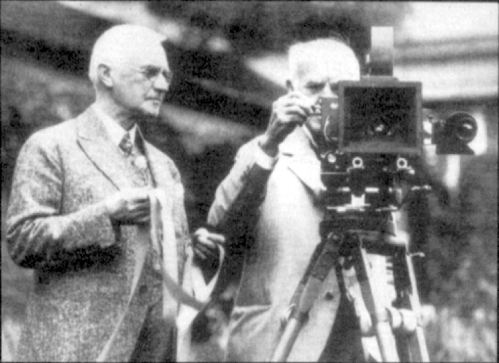 Two men in early 20th-century attire operate a large camera on a tripod. One holds film while the other adjusts the camera.
