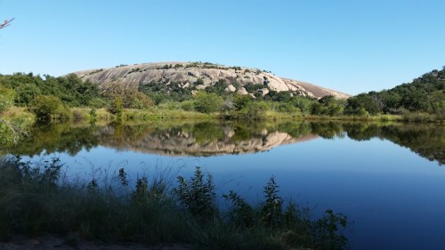 A serene landscape featuring a large rock formation reflected in a calm body of water, surrounded by greenery.