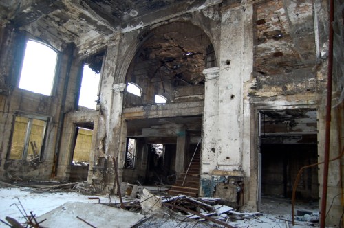 Interior of a dilapidated building with crumbling walls, broken windows, and debris scattered on the floor.