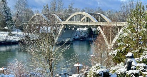 A serene winter scene featuring a snow-covered landscape, a river, and a bridge under a clear blue sky.