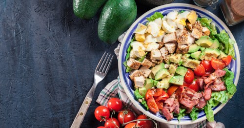 A colorful salad with chicken, avocado, tomatoes, bacon, and hard-boiled eggs, surrounded by fresh ingredients.