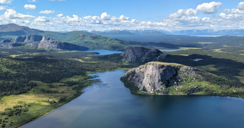 Aerial view of a lush landscape with mountains, lakes, and a clear blue sky dotted with clouds.