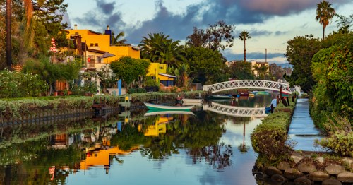 A serene canal scene with colorful houses, a white bridge, and palm trees reflecting in the calm water at sunset.