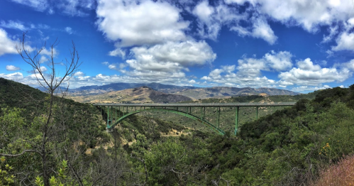 A green bridge spans a valley, surrounded by lush hills and a blue sky with fluffy clouds.