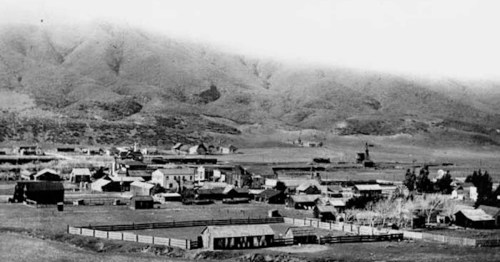 A historic black-and-white photo of a small town surrounded by mountains, featuring wooden buildings and open fields.