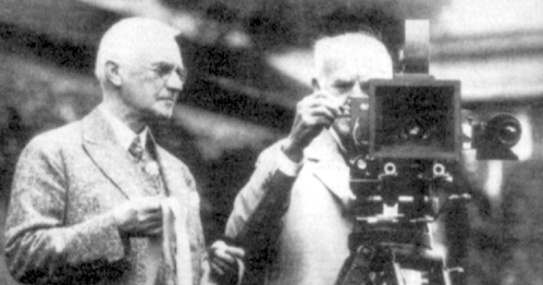 Two men in early 20th-century attire, one operating a large camera, the other observing, outdoors.