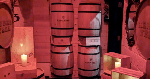 Two wooden wine barrels with labels, surrounded by candlelight and wooden crates in a dimly lit room.
