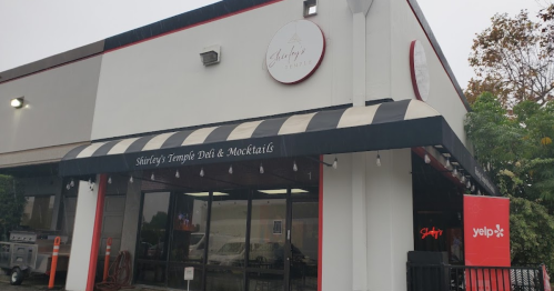 Exterior of Shirley's Temple Deli & Mocktails, featuring a striped awning and a Yelp sign, on a rainy day.