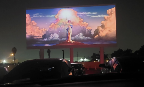 A large illuminated billboard displays a colorful image of a woman standing among clouds at night, with cars in the foreground.
