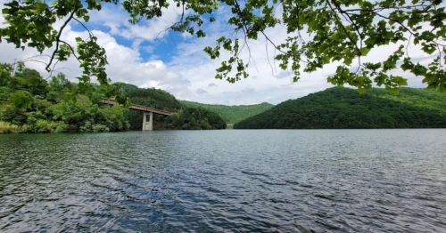 A serene lake surrounded by lush green hills under a partly cloudy sky, with a bridge in the distance.