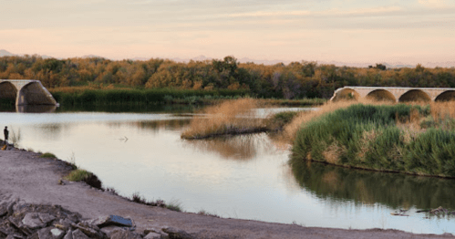 A serene river scene with lush greenery, a stone bridge, and a person fishing along the bank at sunset.