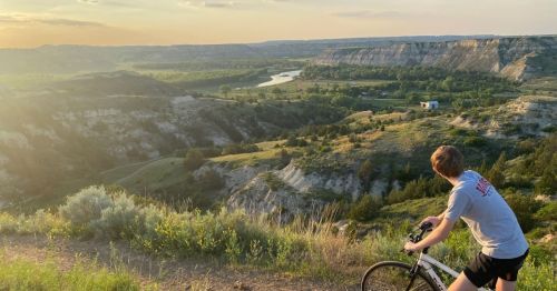 A cyclist overlooks a scenic valley at sunset, with rolling hills and a river in the distance.