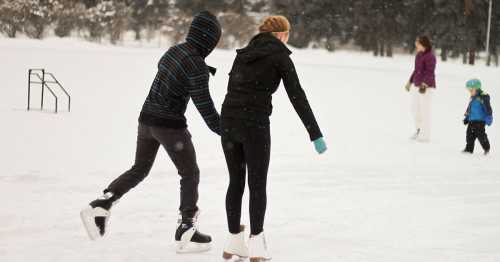 Two people ice skating on a snowy rink, with a child and another person in the background. Snow is falling.