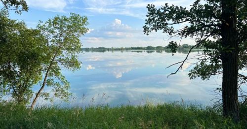 A serene lake reflects the sky and trees, surrounded by lush green grass and gentle clouds.