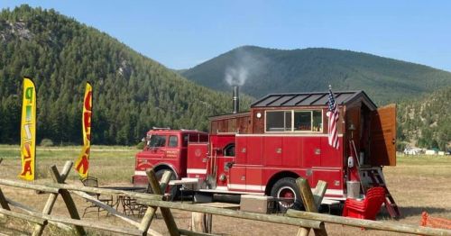 A red fire truck converted into a food truck, parked in a scenic field with mountains in the background.